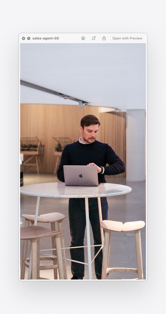 Sales agent standing at a high table using a laptop in a modern, open workspace with wooden stools and background decor.